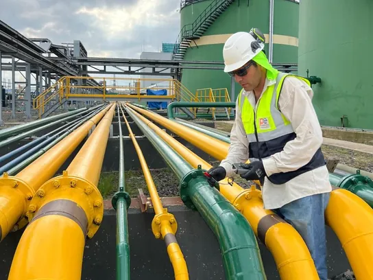 Técnico con chaleco mide sobre las cañerías amarillas y verdes de una planta de combustibles, con los tanques al fondo.