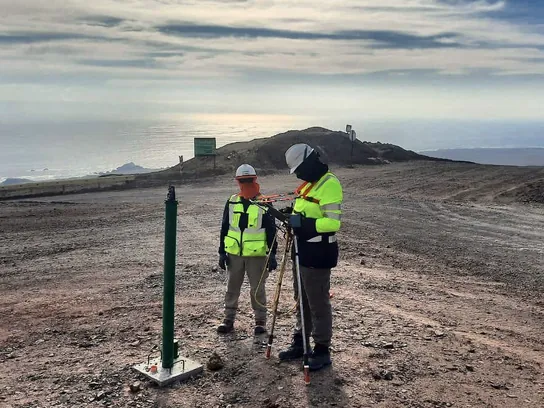 Dos técnicos con casco y chaleco miden junto a una caja de medición, en un camino de servicio frente al mar.