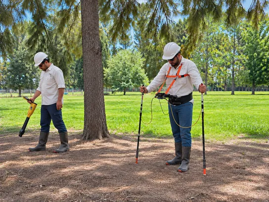 Two technicians in hard hats surveying in the field: one locates the pipeline with a locator and the other runs the DCVG survey with a data logger and two electrodes.