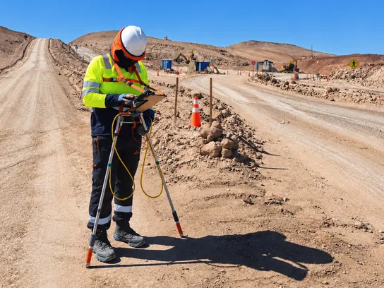 Técnico con EPP completo mide con electrodos y una tablet de registro al costado de un camino de servicio.